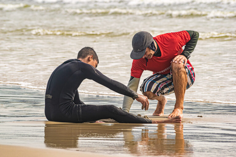 aluno criança da nexus surf school sentado na areia se alongando com o instrutor de surf ernesto na beira de praia da barra da lagoa em florianopolis