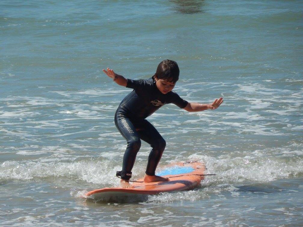 aluno criança da nexus surf school de pé em uma prancha surfando com os braços abertos para tentar se equilibrar na praia da barra da lagoa em florianianópolis